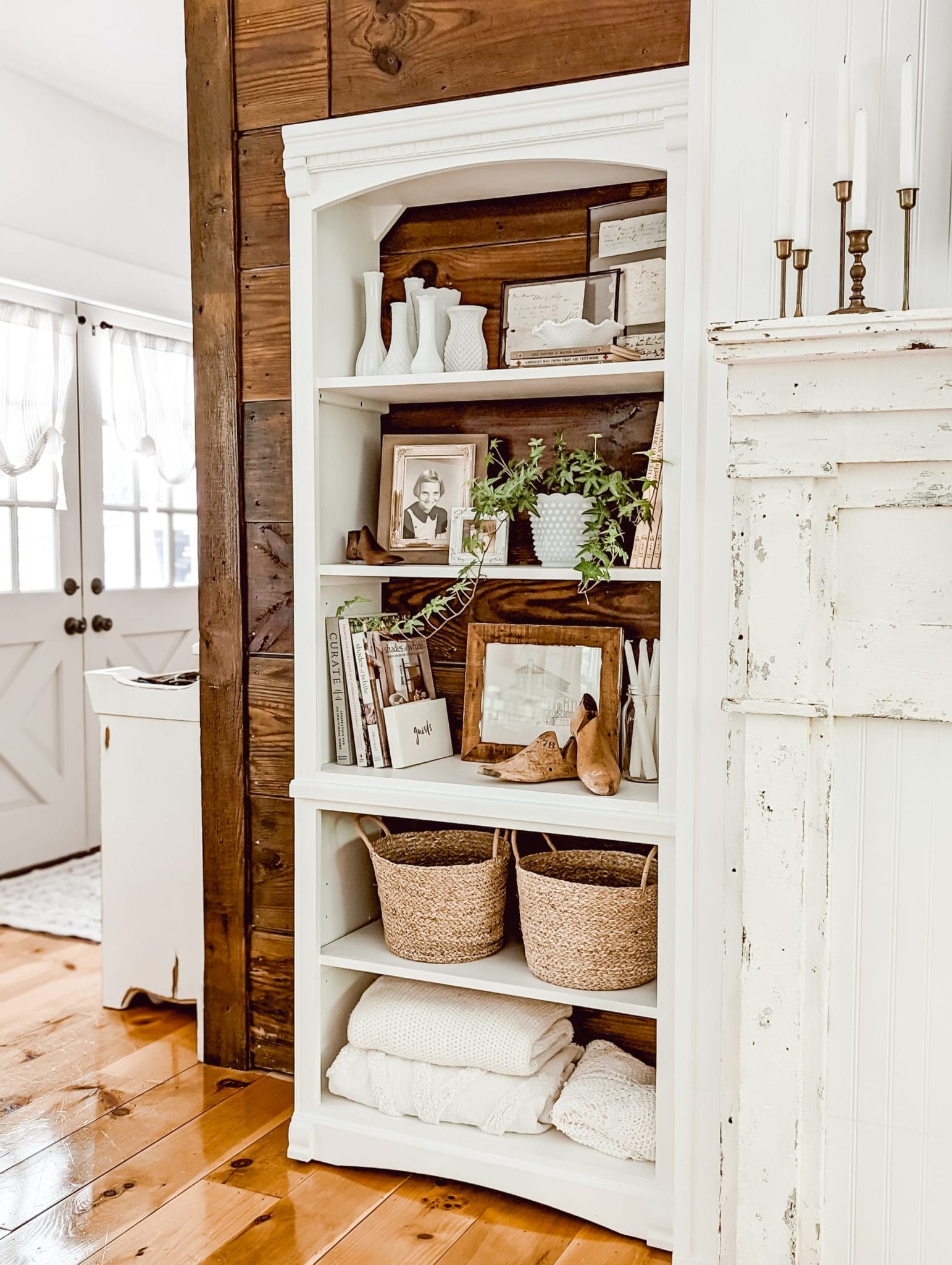 vintage farmhouse and cottage style shelf with framed photos, milk glass vases, wooden shoe molds, a collection of books, baskets, and a houseplant,