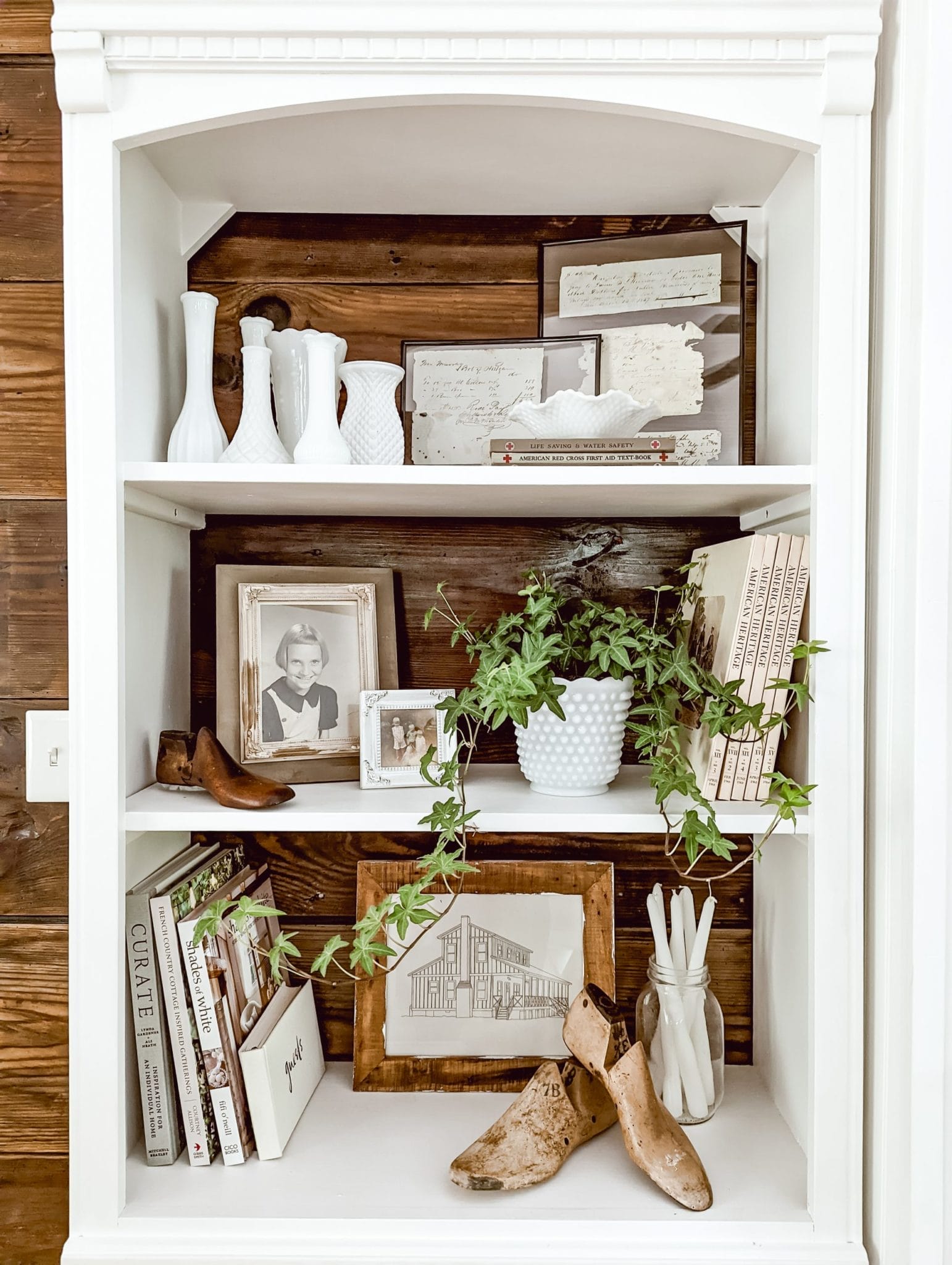 vintage farmhouse and cottage style shelf with framed photos, milk glass vases, a collection of books and an ivy houseplant