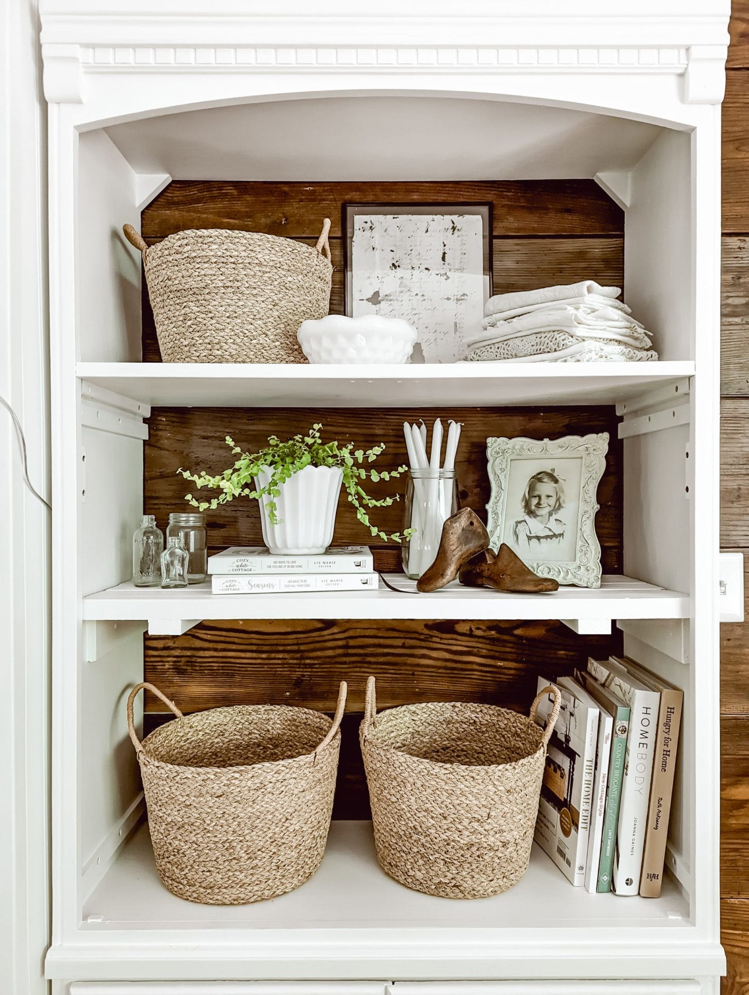 vintage farmhouse and cottage style shelf with framed photos, a stack of doilies, a collection of books, baskets, and a houseplant