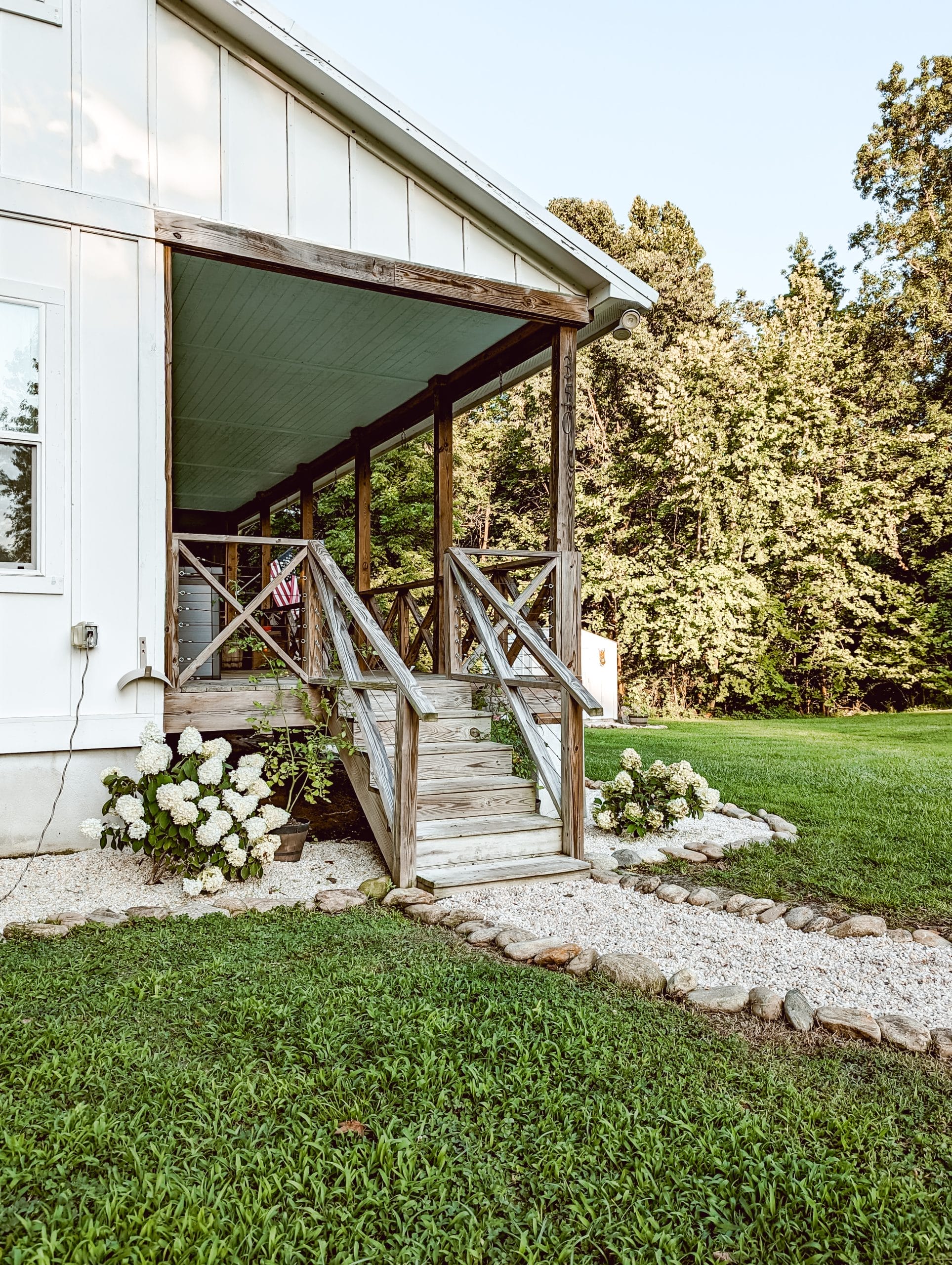 side porch steps leading to a gravel path, and cottage style landscaping with white hydrangeas and pea gravel flower beds around a white farmhouse