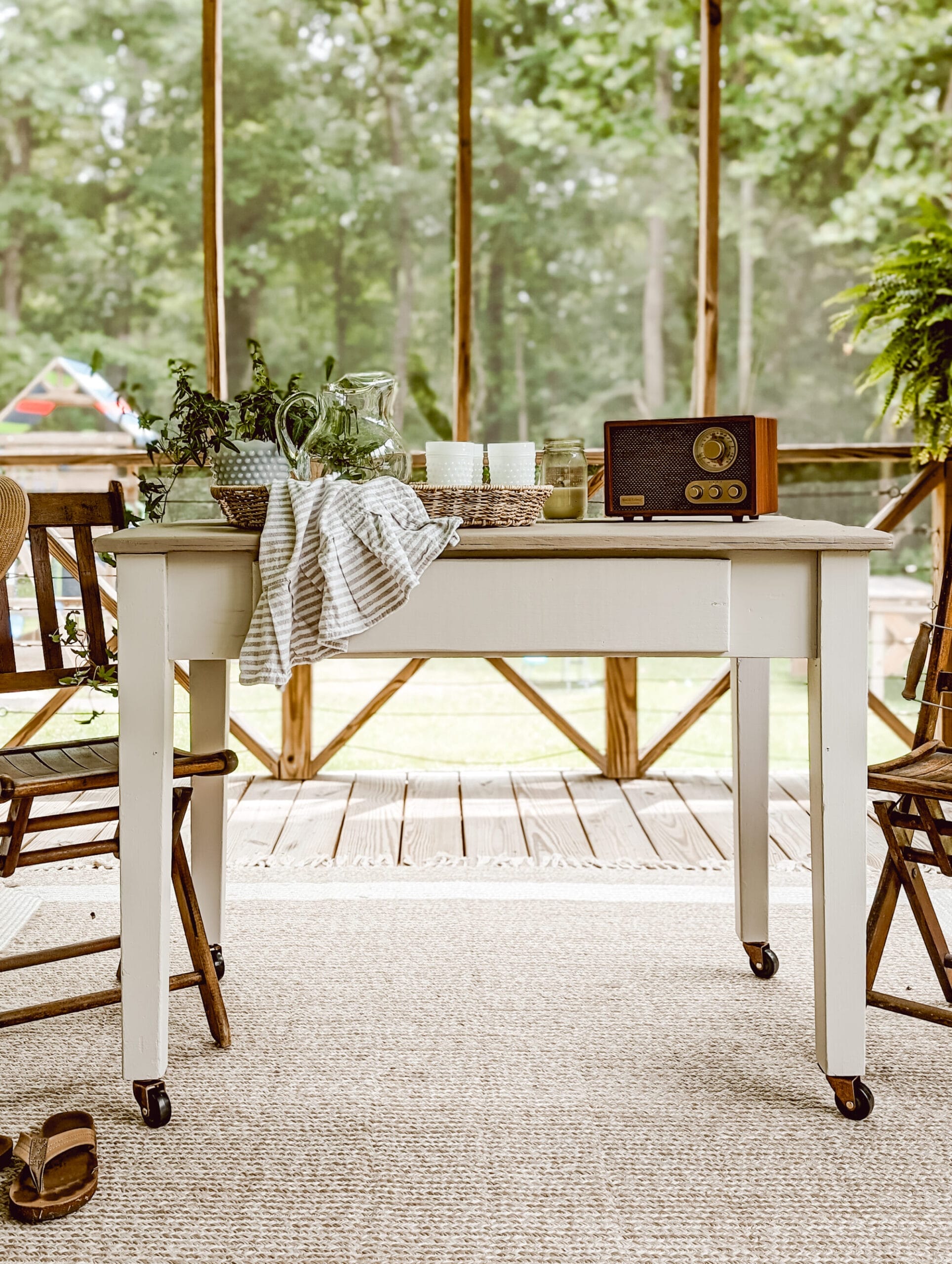painted rolling table on a screened in porch with wood chairs and a wicker tray of vintage water glasses sitting nect to a potted plant and a vintage radio