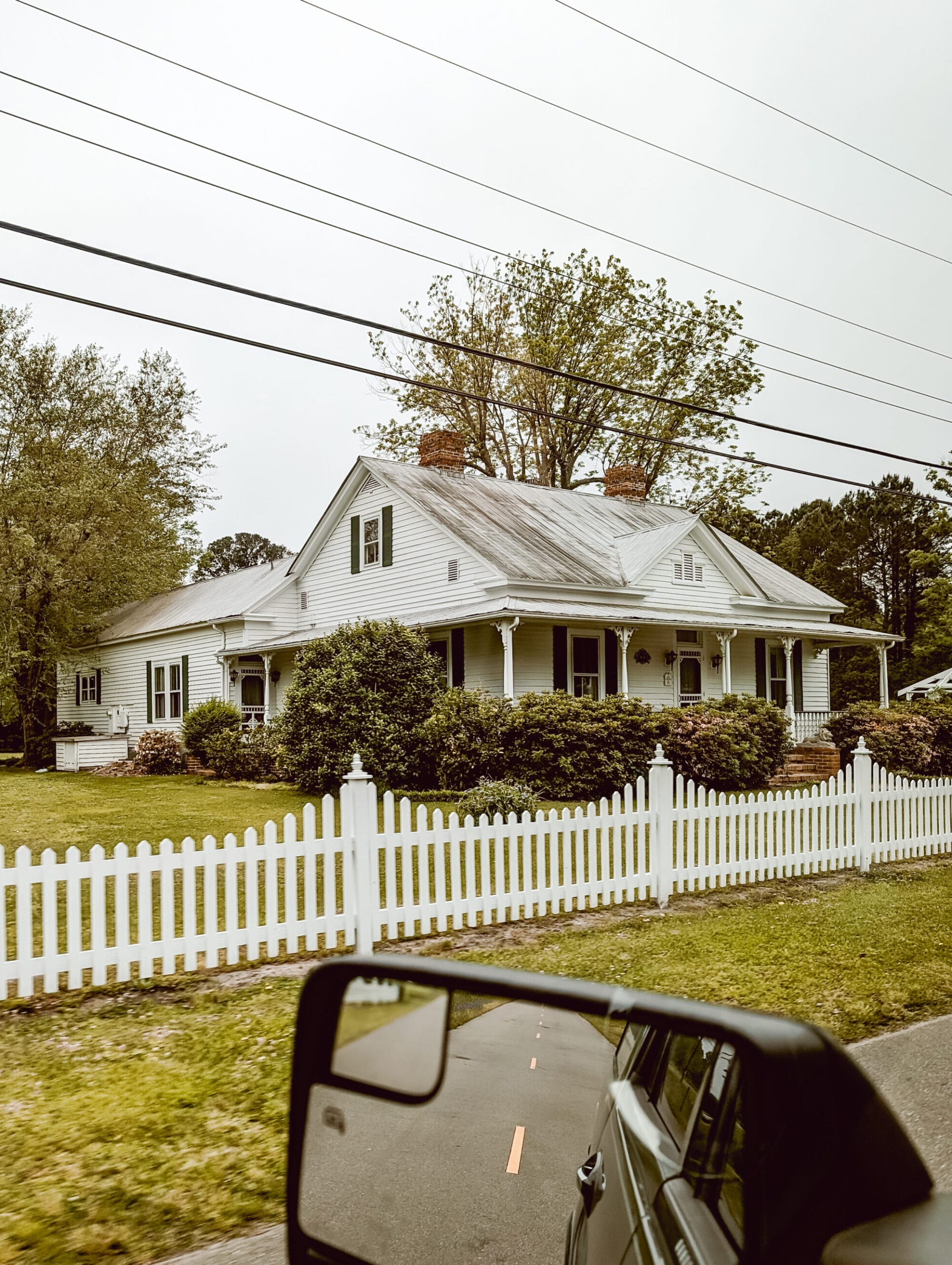 a pretty old house with white siding, green shutters, a metal roof, and pretty corbels on the porch posts with a white picket fence along the road
