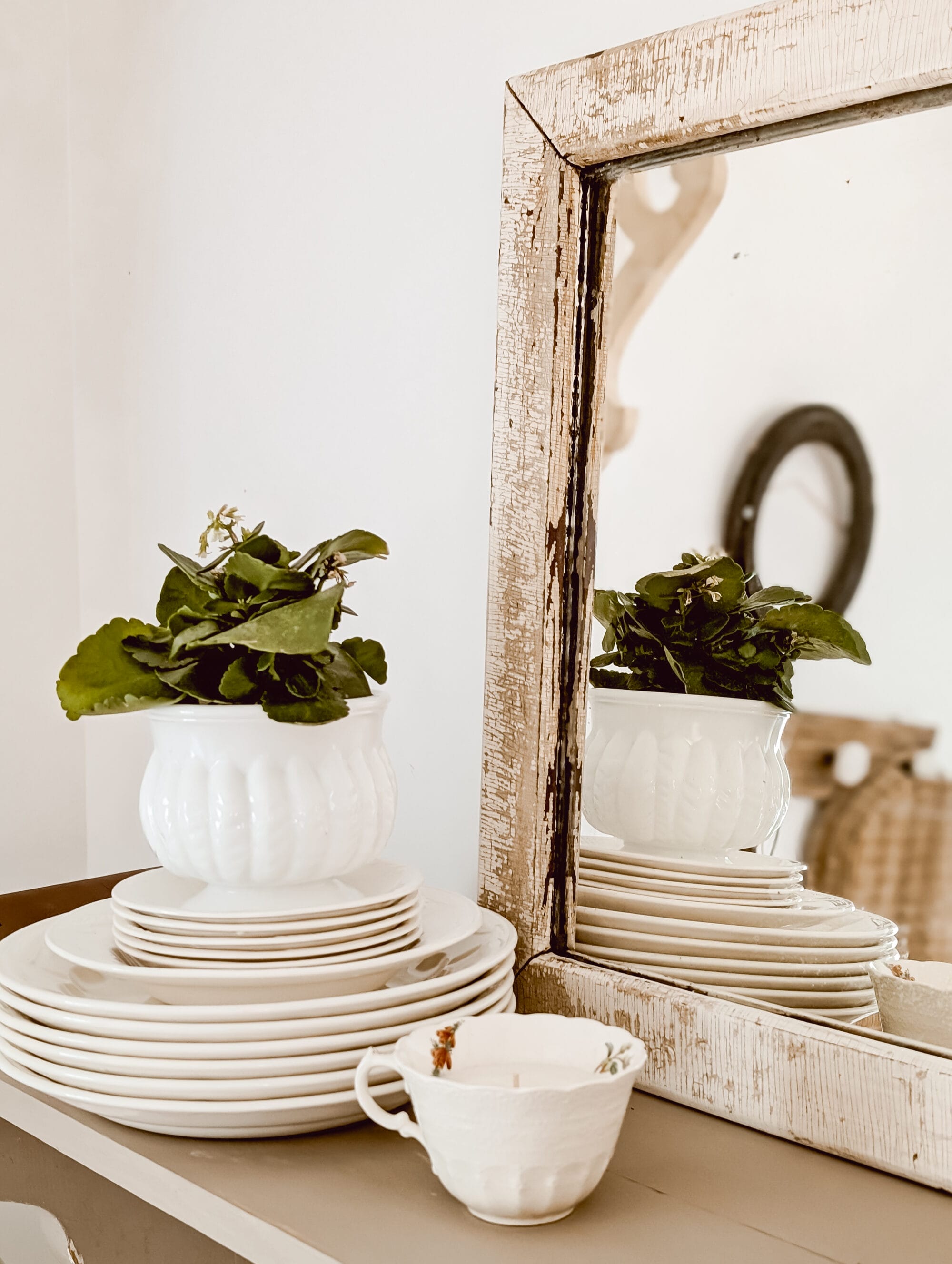 vintage white dishes stacked and topped with a milk glass planter next to a chippy antique mirror