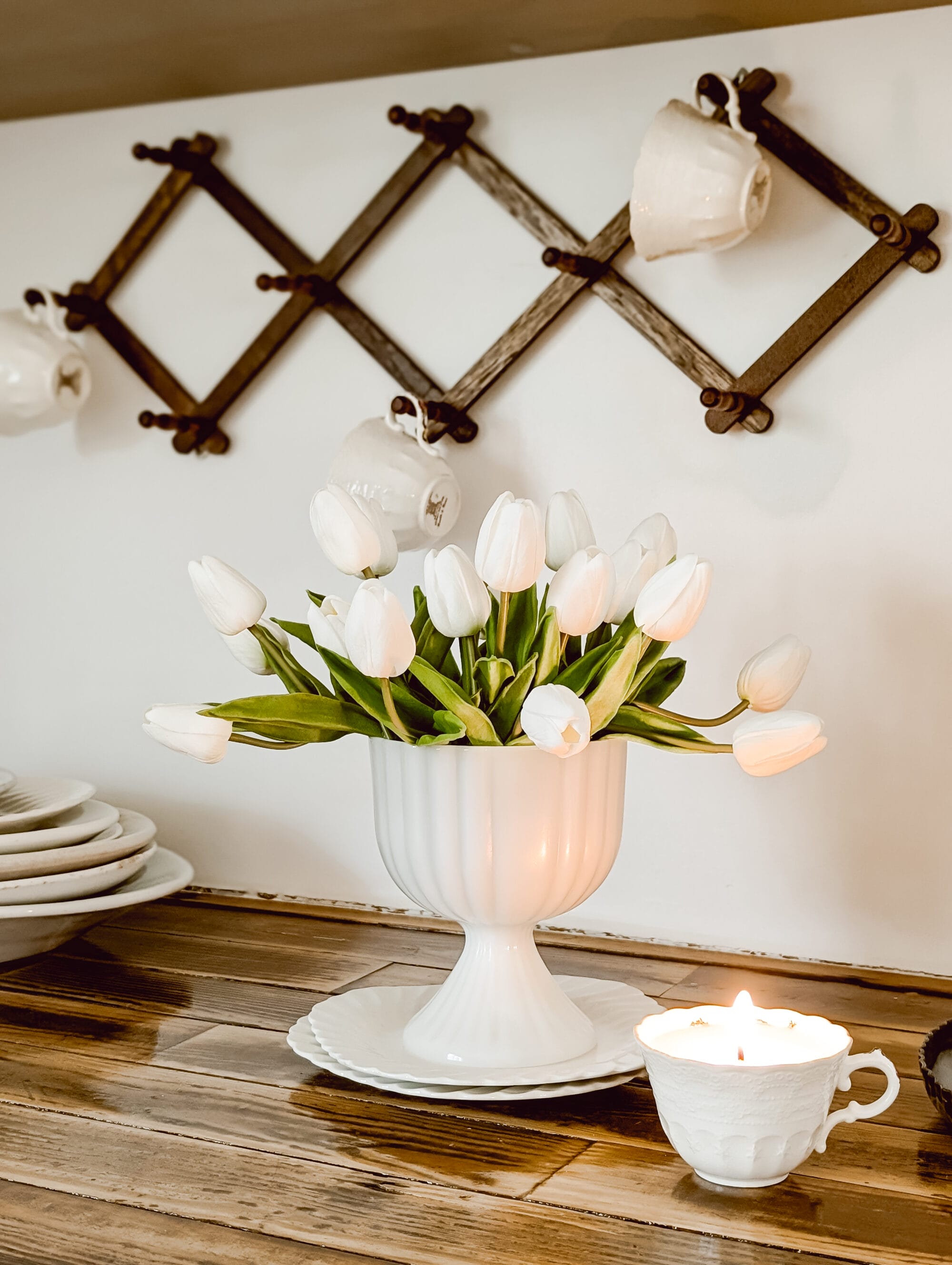 real touch white tulips arranged in a milk glass pedestal planter and set on top of two pretty scalloped dishes next to a vintage teacup candle