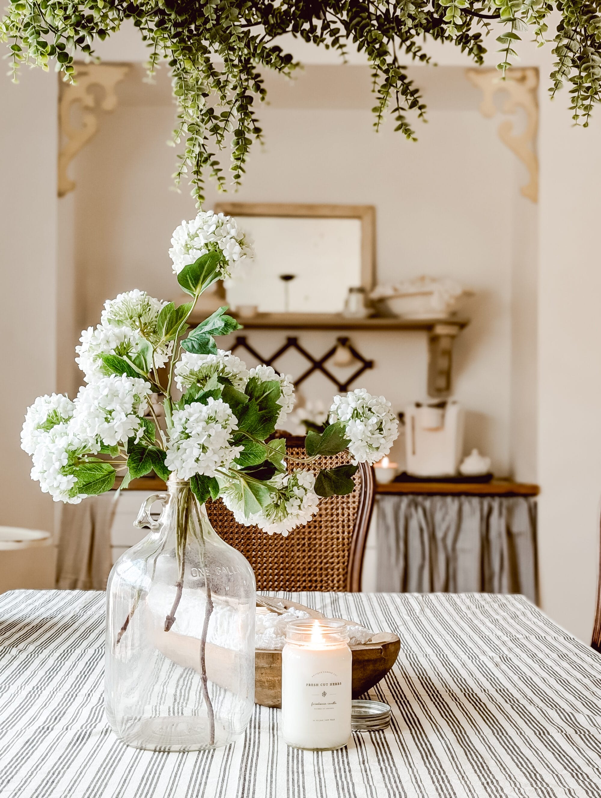 old glass gallon jug with faux white hydrangea stems next to a Fresh Cut Herbs candle from Antique Candle Co atop a white and black striped tablecloth