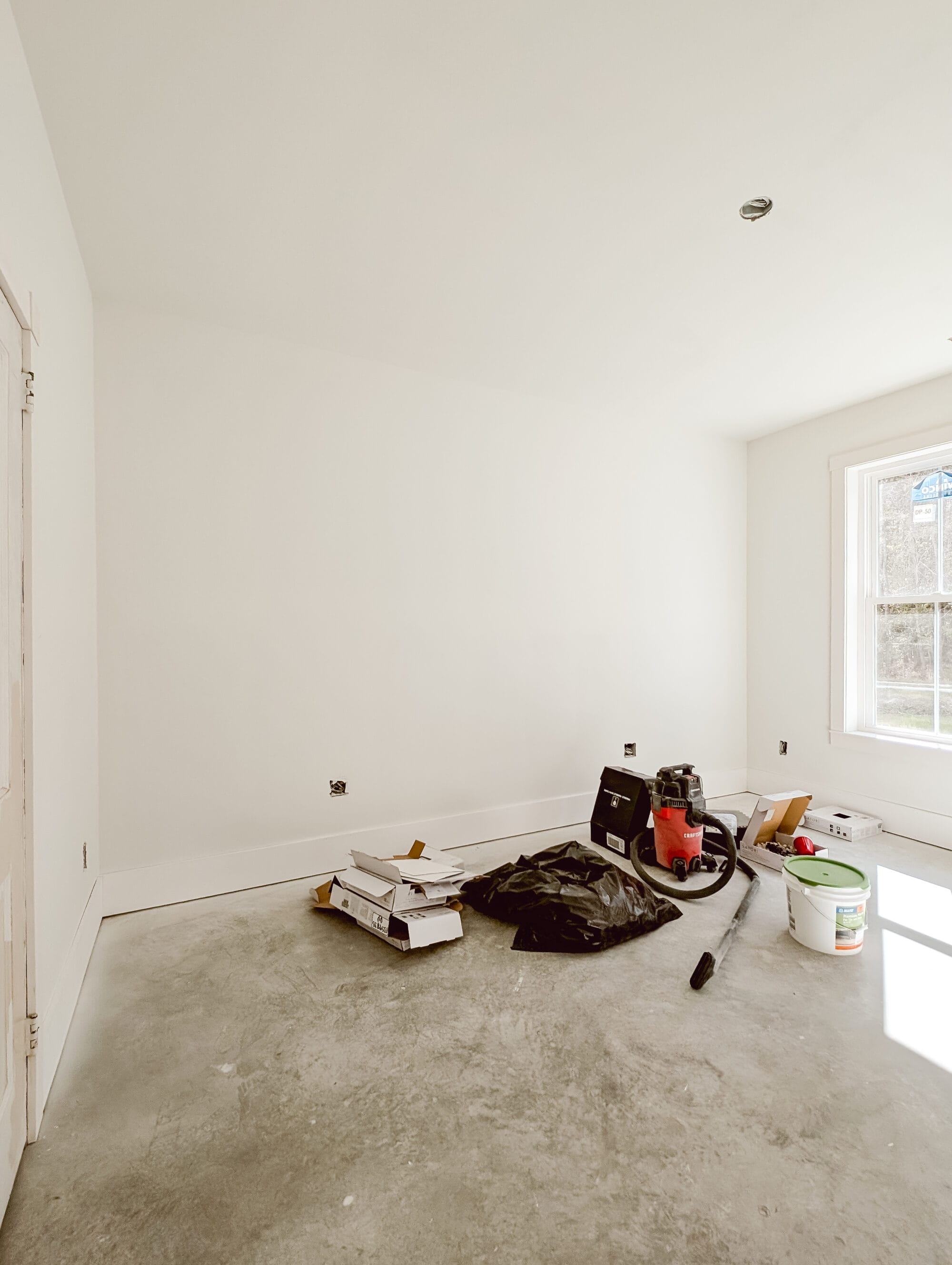 bed wall in our unfinished primary bedroom with primed walls and concrete subfloors and building supplies on the floor