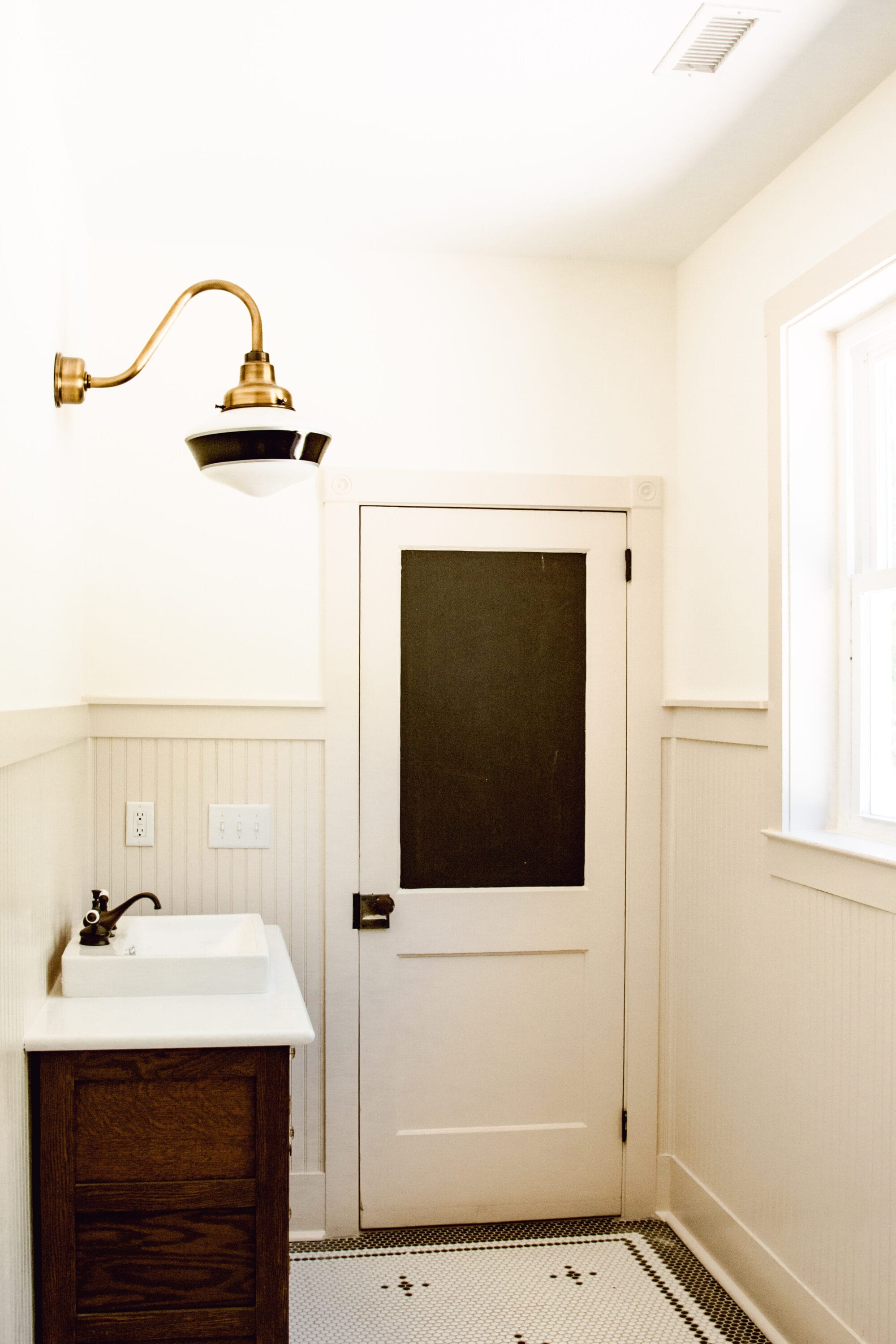 primary bathroom built new but designed to look like an older home's bathroom with antique door, antique vanity, vintage schoolhouse light, and black and white penny tile floors