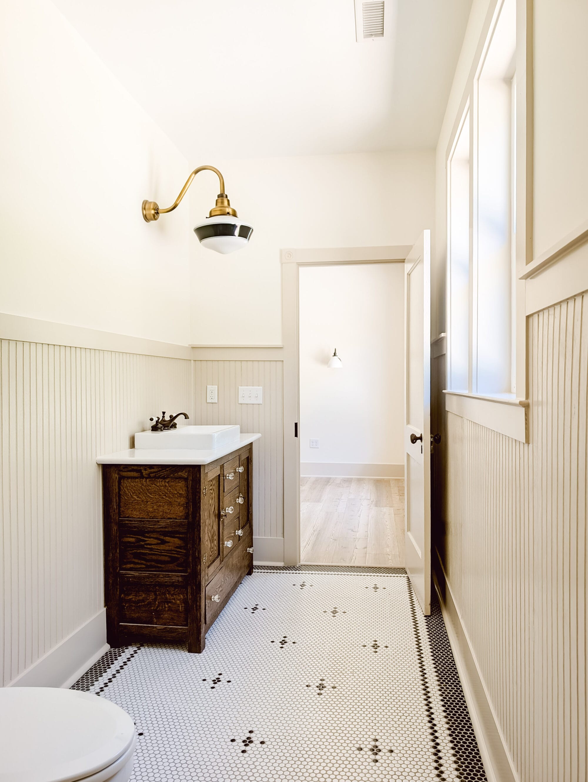 vintage cottage bathroom with beadboard paneling, black and white penny tile floors, a gooseneck schoolhouse vanity light, and an antique vanity