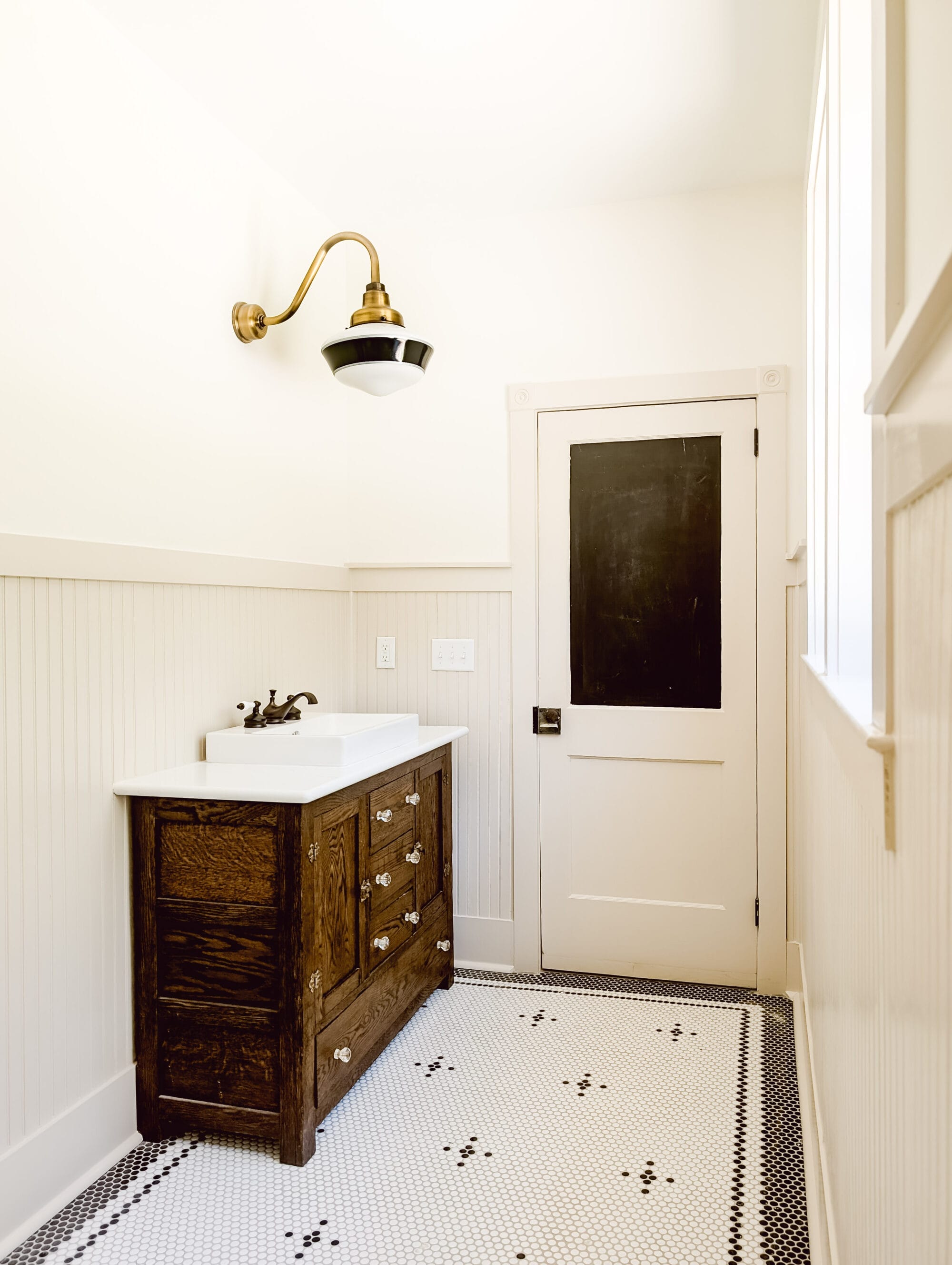 vintage cottage bathroom with black and white penny tile floors, a gooseneck schoolhouse vanity light, and an antique vanity and salvaged door